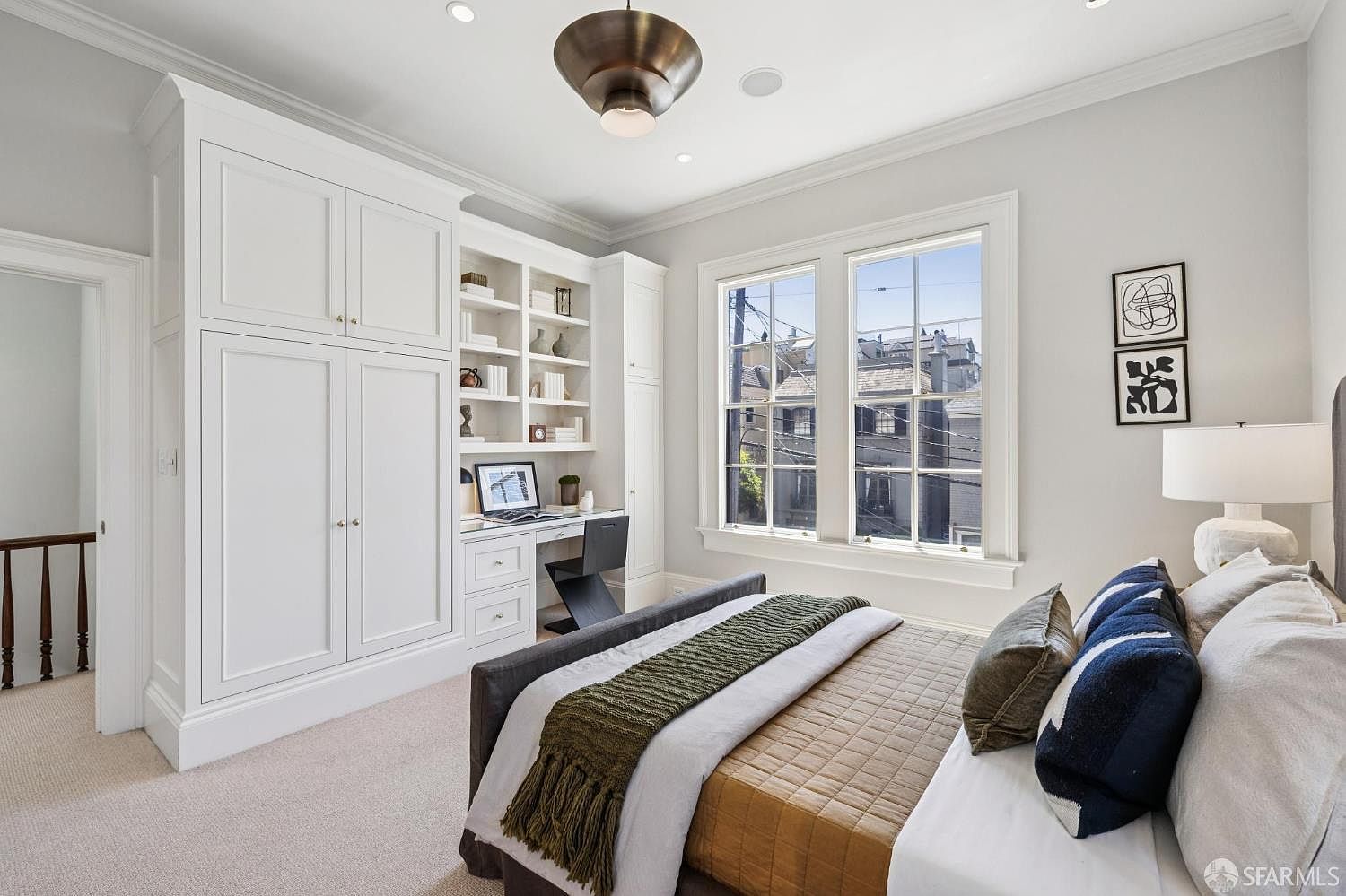 This bright and airy bedroom features a built-in desk and storage unit, maximizing the utility of the space. A queen-sized bed with a dark frame and layered bedding serves as the focal point, positioned near large windows that provide natural light and an urban view. The room is finished with neutral walls and carpet, creating a clean, versatile aesthetic suitable for a guest room or home office.