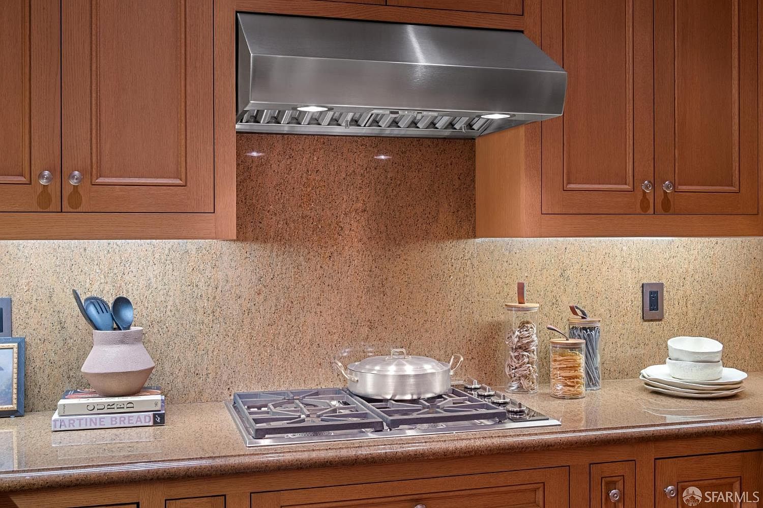 This kitchen features warm-toned wood cabinetry and a stone backsplash that complements the matching stone countertop. A stainless steel gas cooktop with a matching range hood serves as the focal point, while decorative glass canisters and a stack of bowls add a lived-in, functional touch. The perspective is a straight-on, eye-level shot that highlights the clean, organized workspace and traditional aesthetic.