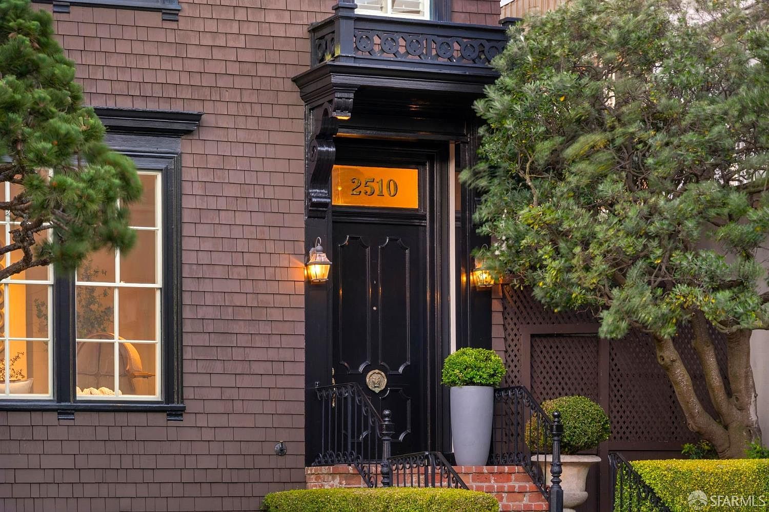 This elegant front entryway features a dark, paneled door framed by a decorative black architectural surround and a prominent gold house number plaque reading 2510. The exterior is clad in dark brown shingles, complemented by black window frames and a manicured hedge in the foreground. A potted plant sits beside the steps, adding a touch of greenery to the sophisticated, classic facade.