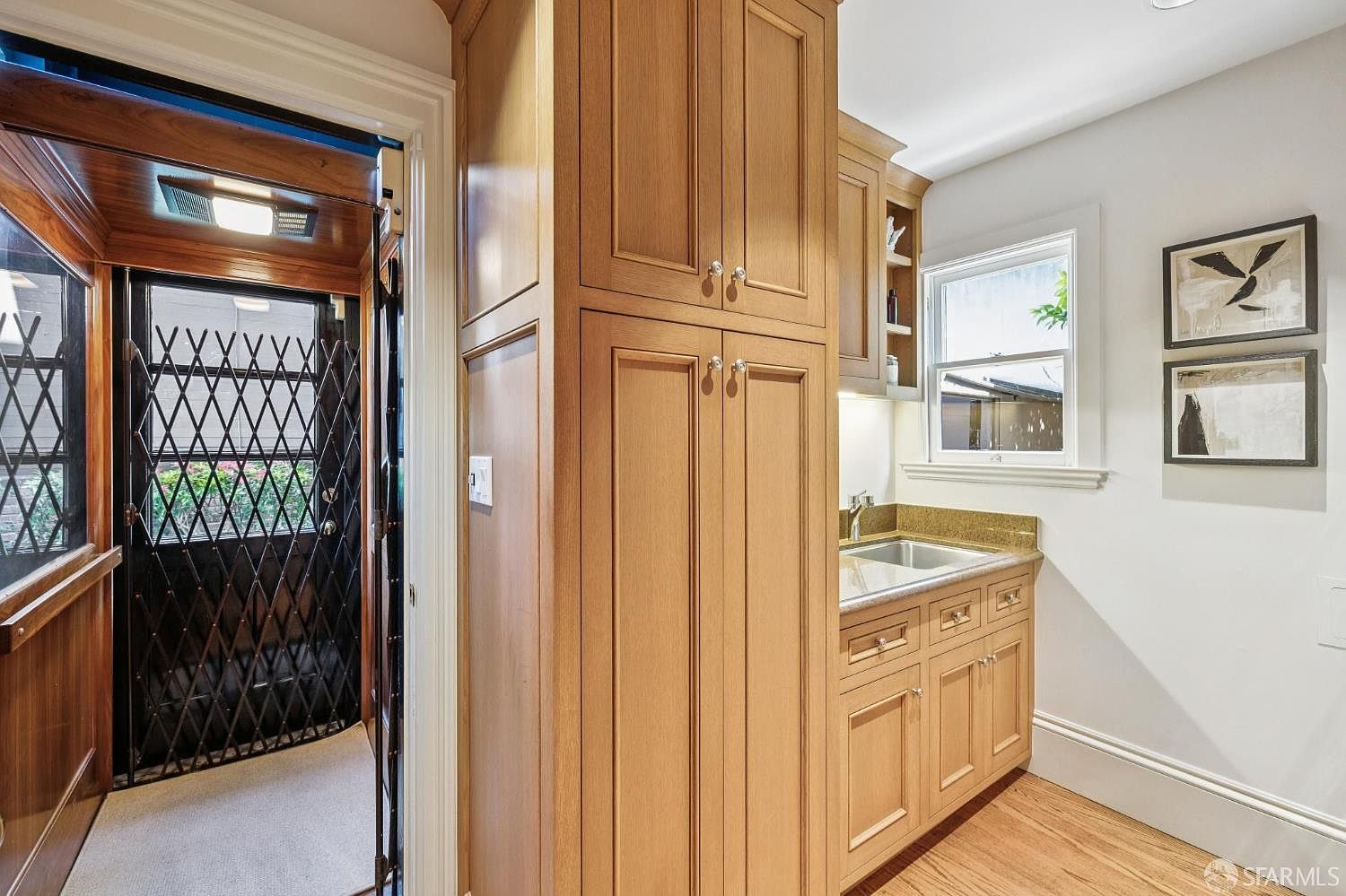 This interior view captures a functional wet bar or butler's pantry area featuring light wood cabinetry and a stone countertop with an integrated sink. To the left, a doorway with a decorative metal security gate leads to an exterior space, while the right wall is adorned with two framed pieces of art. The perspective highlights the transition between the interior utility space and the outdoor entry, showcasing a clean and organized aesthetic.