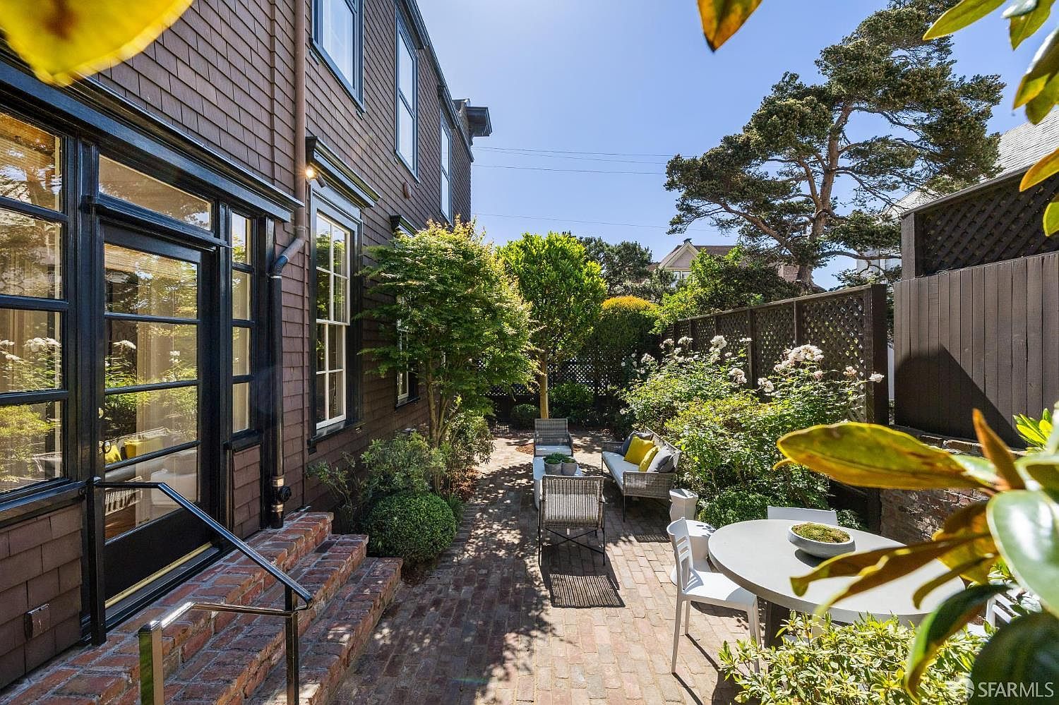 This inviting outdoor patio features a classic brick-paved floor and is framed by a dark-stained wood-shingled house exterior with black-trimmed windows. The space is furnished with comfortable seating, including a sofa and chairs, and a round dining table, all nestled among lush greenery and mature trees. The perspective is from a slightly elevated position, looking out over the private, enclosed garden area that offers a serene and secluded atmosphere.