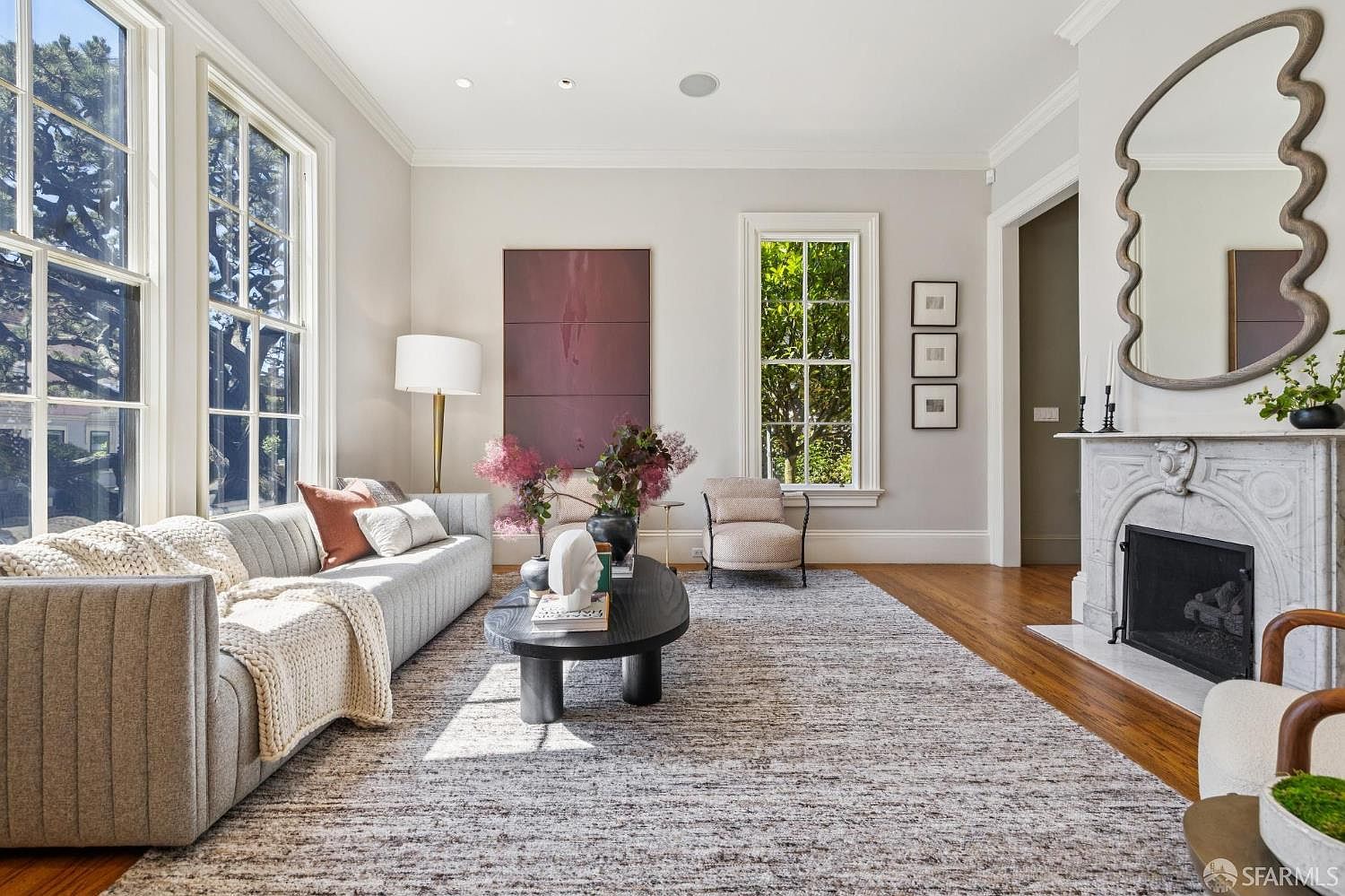 This bright and airy living room features a neutral color palette, tall windows that flood the space with natural light, and warm wood flooring. A plush gray sofa with textured throws sits opposite a stone fireplace mantel adorned with a large, uniquely shaped mirror. The room is accented by a dark oval coffee table, a modern floor lamp, and a large piece of abstract art, creating a sophisticated and inviting atmosphere.