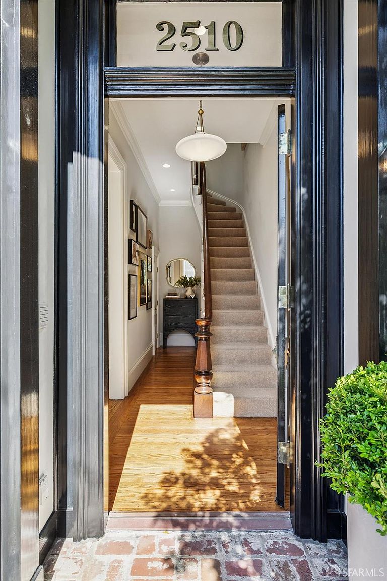 This inviting entryway features a classic black-framed doorway that opens into a bright, welcoming foyer. Inside, a staircase with a wooden banister leads upward, complemented by warm wood flooring and a gallery wall of framed art. The perspective from the brick-paved threshold offers a clear, cinematic view into the home's elegant interior.