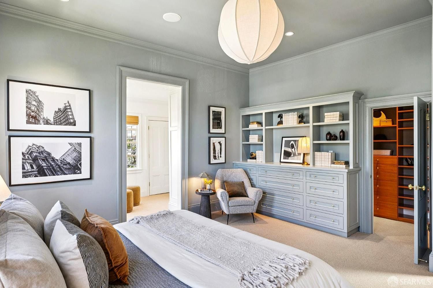 This elegant primary bedroom features a soft blue-gray color palette, plush carpeting, and a large, textured paper pendant light fixture. A custom built-in unit provides ample storage with drawers and open shelving, complemented by a cozy armchair and a small side table. The room flows into an adjacent walk-in closet and a secondary hallway, creating a sophisticated and well-organized living space.