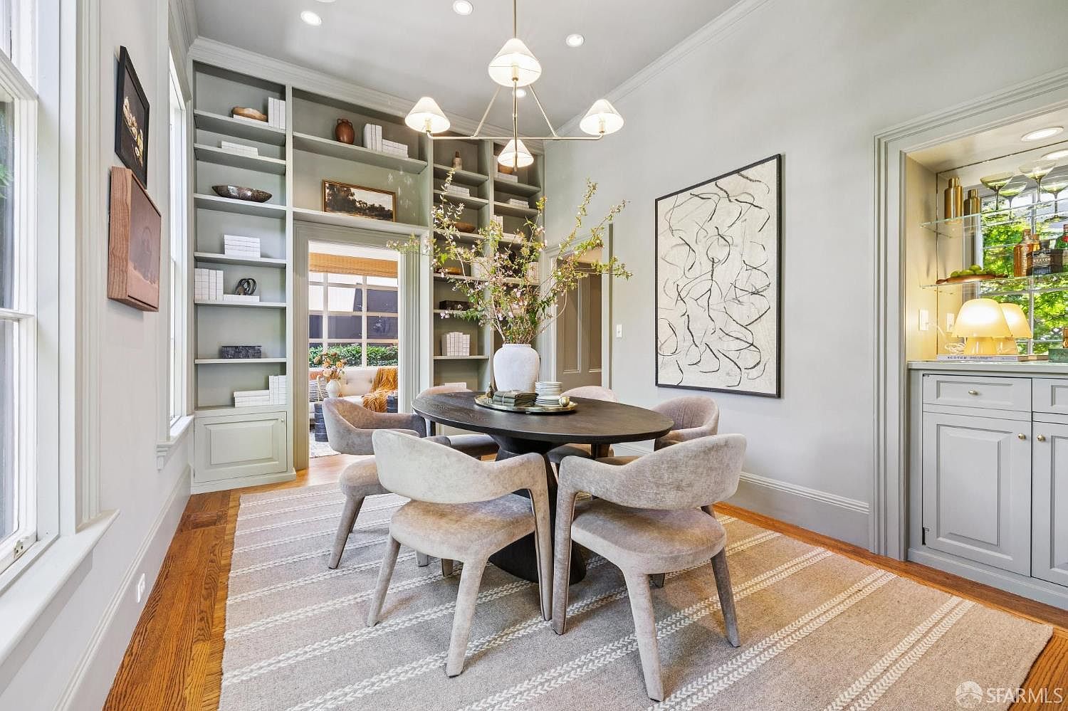 This elegant dining room features a dark oval table surrounded by four upholstered chairs, set atop a textured area rug on hardwood floors. Built-in shelving units flank a doorway leading to an adjacent room, while a modern chandelier hangs above a central floral arrangement. To the right, a built-in bar area with glass shelving and a mirrored back adds a sophisticated touch, complemented by a large abstract art piece on the wall.