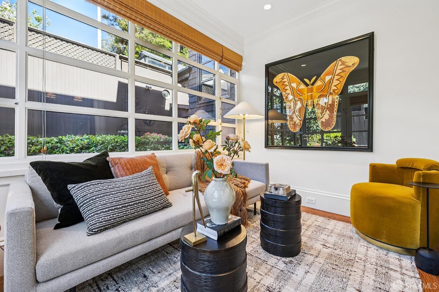 This bright and inviting living room features a light gray sofa adorned with textured throw pillows and a central vase of flowers. A large, framed butterfly art piece serves as a striking focal point on the wall, complemented by a vibrant mustard-yellow armchair and two dark, cylindrical side tables. The space is illuminated by natural light streaming through a large grid-patterned window wall, creating a warm and sophisticated atmosphere.