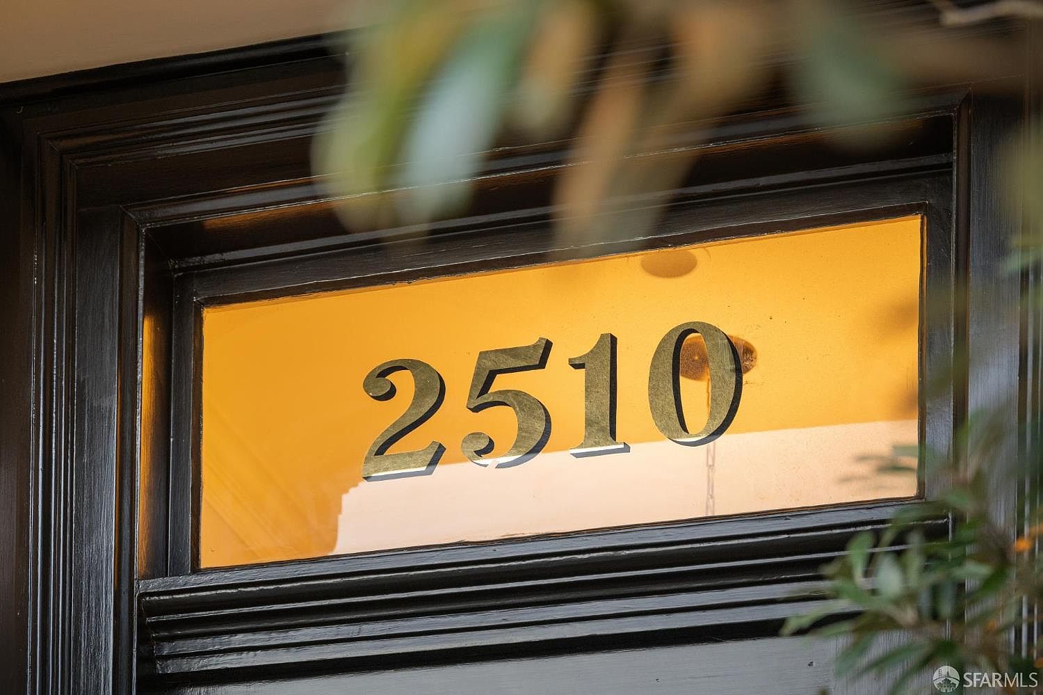 This close-up shot captures a classic transom window above a front door, featuring the house number '2510' in bold, dark, dimensional lettering. The glass is backlit with a warm, inviting glow, framed by a dark, polished wood trim that adds a touch of traditional elegance. The perspective is centered and eye-level, highlighting the architectural detail of the entryway.