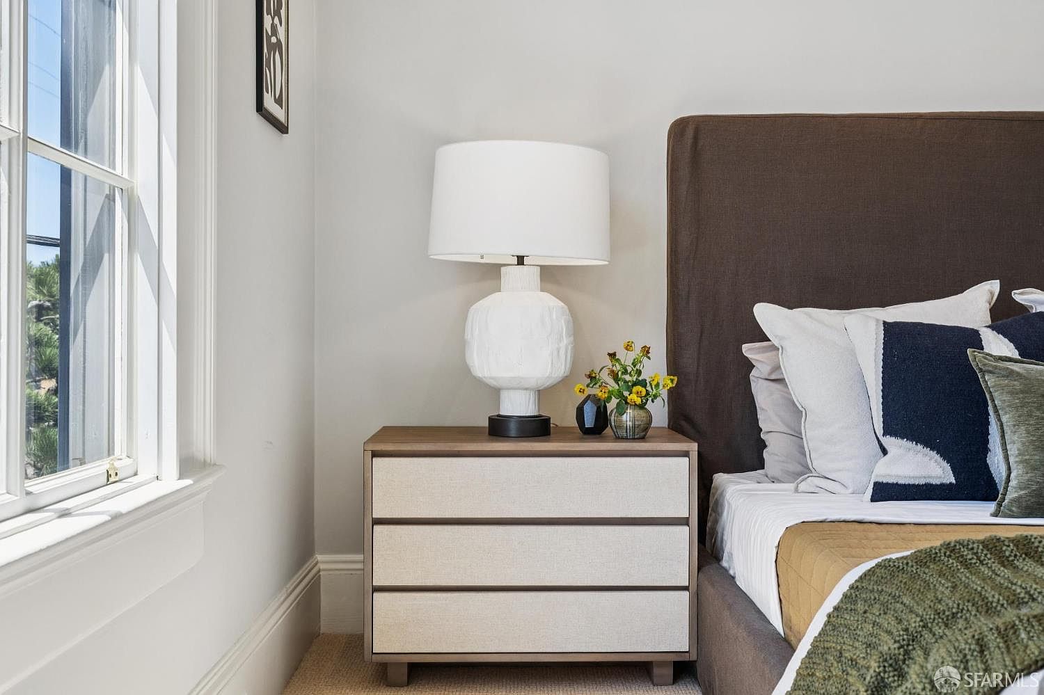 This close-up shot of a bedroom corner features a modern nightstand with light-colored drawer fronts and a textured white table lamp. To the right, a portion of a plush, dark-toned upholstered headboard is visible, accented by layered pillows in neutral and navy tones. A window to the left allows natural light to brighten the space, creating a serene and inviting atmosphere.