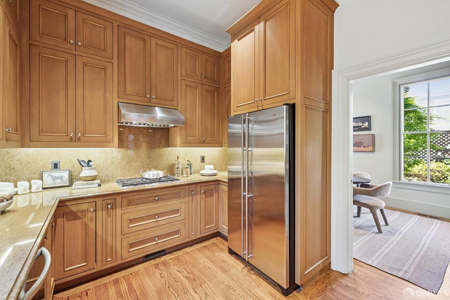 This kitchen features warm-toned wood cabinetry, a stainless steel refrigerator, and a gas cooktop with a metal range hood. The countertops are made of a speckled stone material, and the space is illuminated by under-cabinet lighting. A doorway leads into an adjacent dining area, providing a view of a bright room with large windows and a patterned rug.