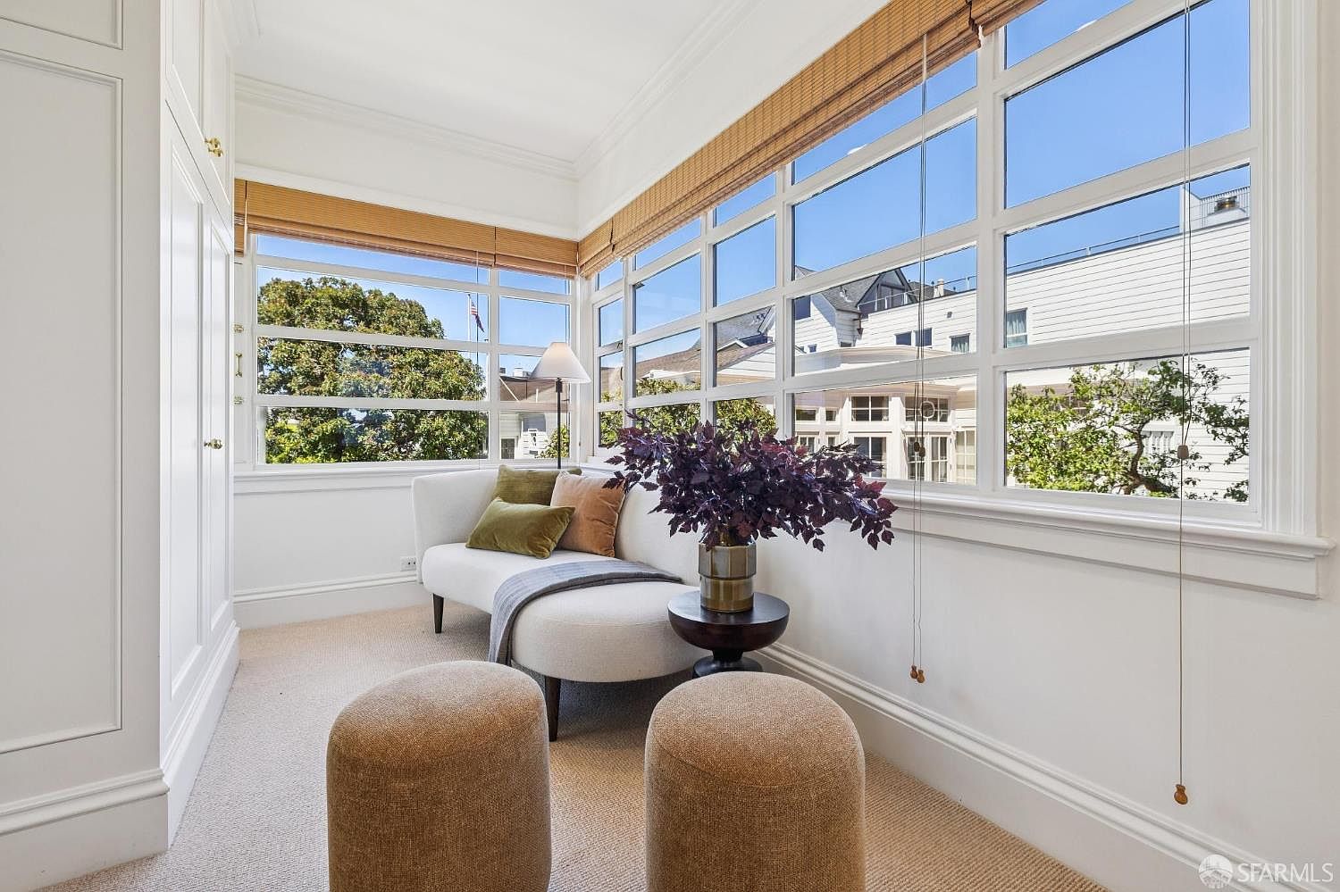 This bright and airy sunroom features expansive windows that wrap around the space, offering abundant natural light and views of the surrounding neighborhood. The room is furnished with a sleek, curved sofa accented with earth-toned pillows, a small round side table topped with a lush plant, and two textured ottomans. The neutral color palette and clean lines create a serene, inviting atmosphere perfect for relaxation.
