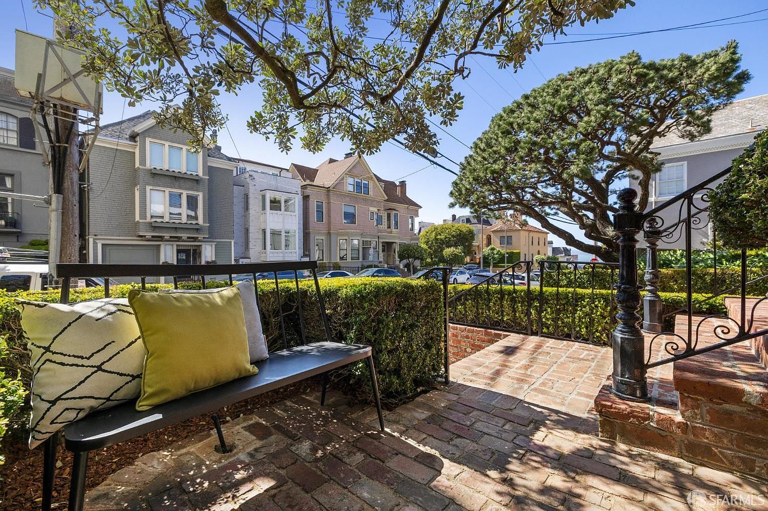 This inviting outdoor patio space features a brick-paved surface and a black metal bench adorned with decorative pillows, set against a backdrop of manicured hedges. The perspective looks out from the elevated terrace toward a quiet residential street lined with classic multi-story homes and a large, mature pine tree. The scene captures a peaceful, sunny atmosphere, highlighting the home's curb appeal and private outdoor seating area.