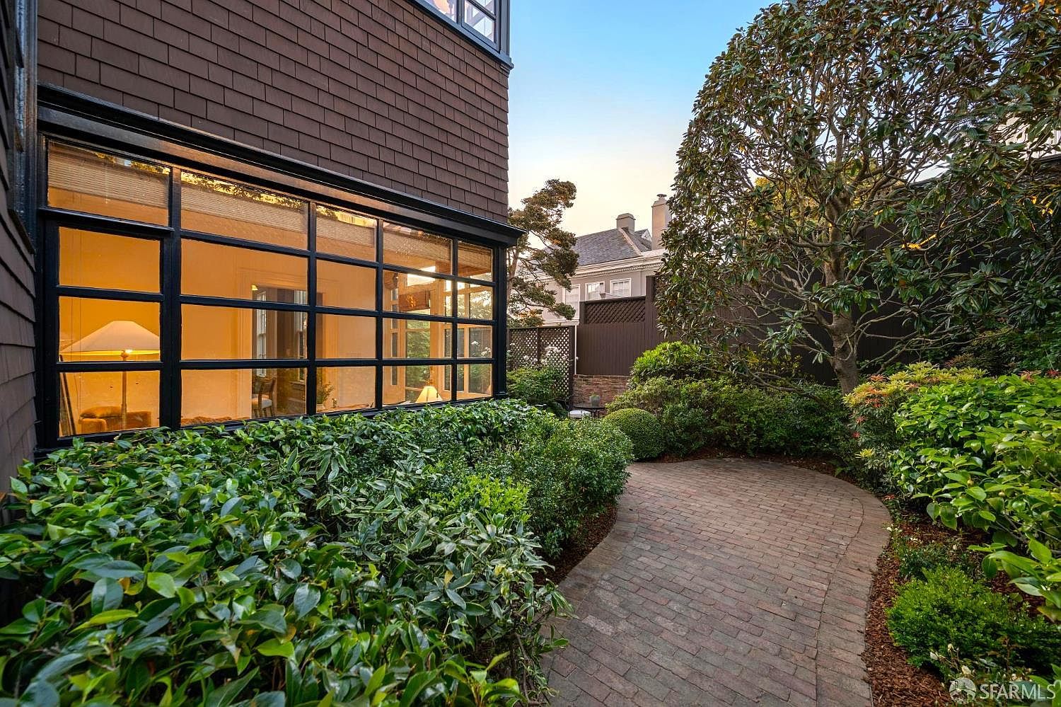 This exterior view captures a lush, private garden space featuring a winding brick pathway that leads toward the back of the property. A large, multi-paned window wall on the side of the dark-shingled house provides a glimpse into the interior living space, creating a seamless connection between the home and the outdoors. The scene is framed by mature trees and dense, manicured shrubbery, offering a serene and secluded atmosphere.