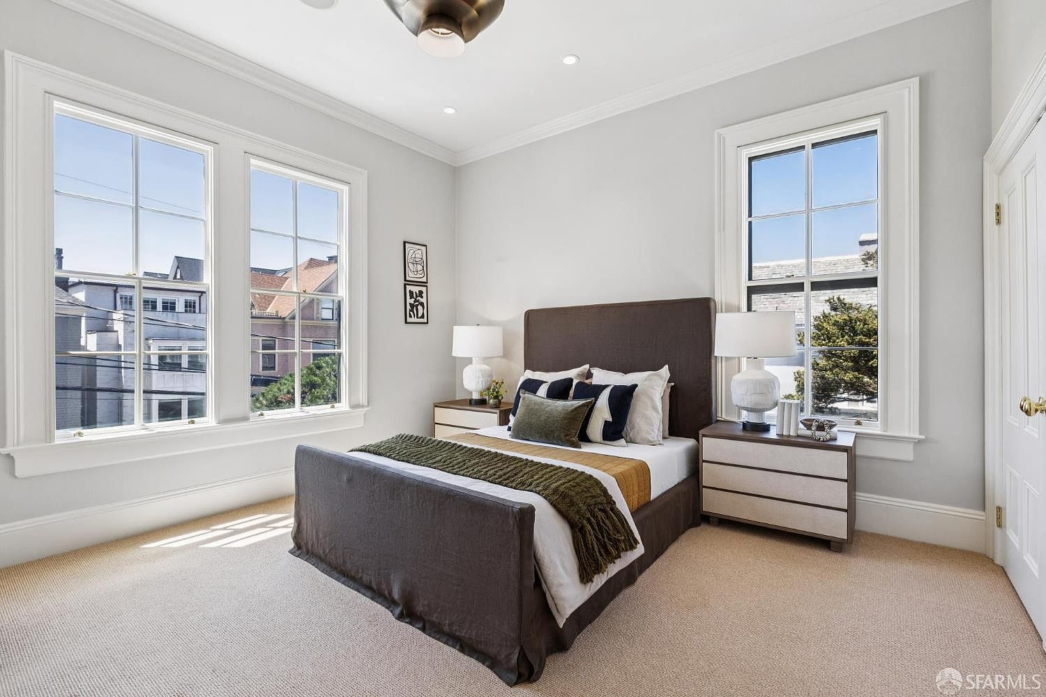 This bright and airy bedroom features a neutral color palette with a dark upholstered bed frame as the central focal point. The room is illuminated by natural light streaming through three large windows, highlighting the light-toned carpeting and soft gray walls. Two matching nightstands with white lamps flank the bed, creating a balanced and inviting atmosphere.