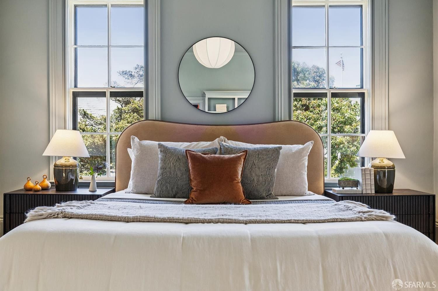 This primary bedroom features a symmetrical layout centered around a plush, curved headboard upholstered in a soft, muted tone. Flanking the bed are two dark, slatted-front nightstands topped with classic table lamps, while a circular mirror hangs between two tall windows that provide ample natural light. The room is decorated with a layered bedding ensemble, including a textured throw blanket, creating a serene and sophisticated atmosphere.