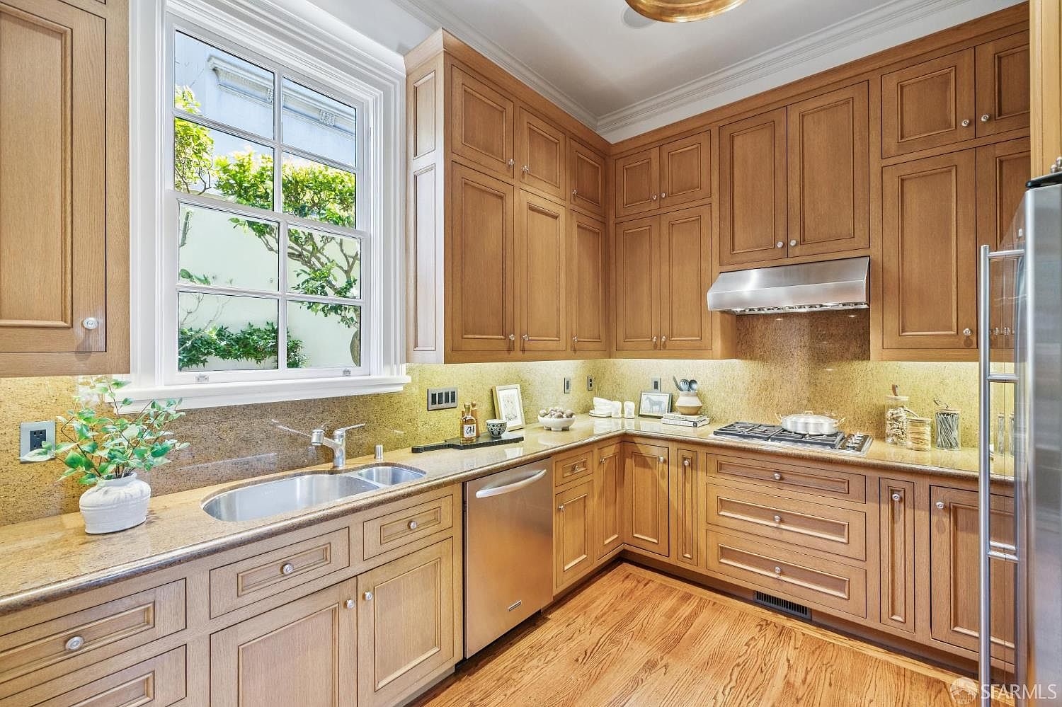 This well-appointed kitchen features warm-toned wood cabinetry, stone countertops, and a suite of stainless steel appliances. A large window above the sink provides natural light and a view of greenery, while under-cabinet lighting highlights the workspace. The space is designed with a classic, functional layout that includes a gas cooktop and a built-in dishwasher.