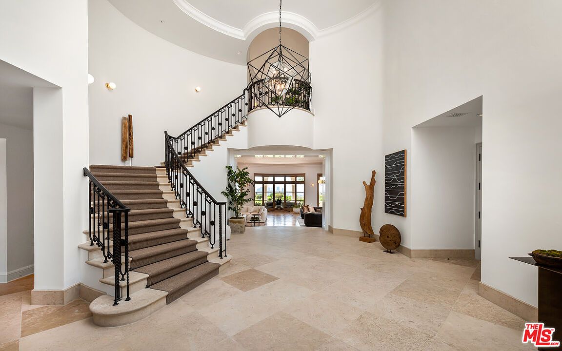 This grand foyer features a sweeping staircase with wrought iron railings and carpeted steps, leading to an upper level. The high ceilings are accentuated by a modern chandelier, and the travertine flooring adds a touch of elegance. The space is bright and airy, with views into the living areas beyond.