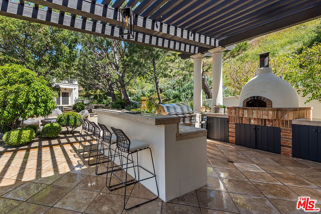 This image showcases an outdoor kitchen and patio area, featuring a built-in grill, a pizza oven constructed with brick and stucco, and a bar counter with seating. The patio is paved with stone tiles, and a pergola provides shade, creating an inviting space for outdoor entertaining. Lush greenery surrounds the area, enhancing the natural ambiance.