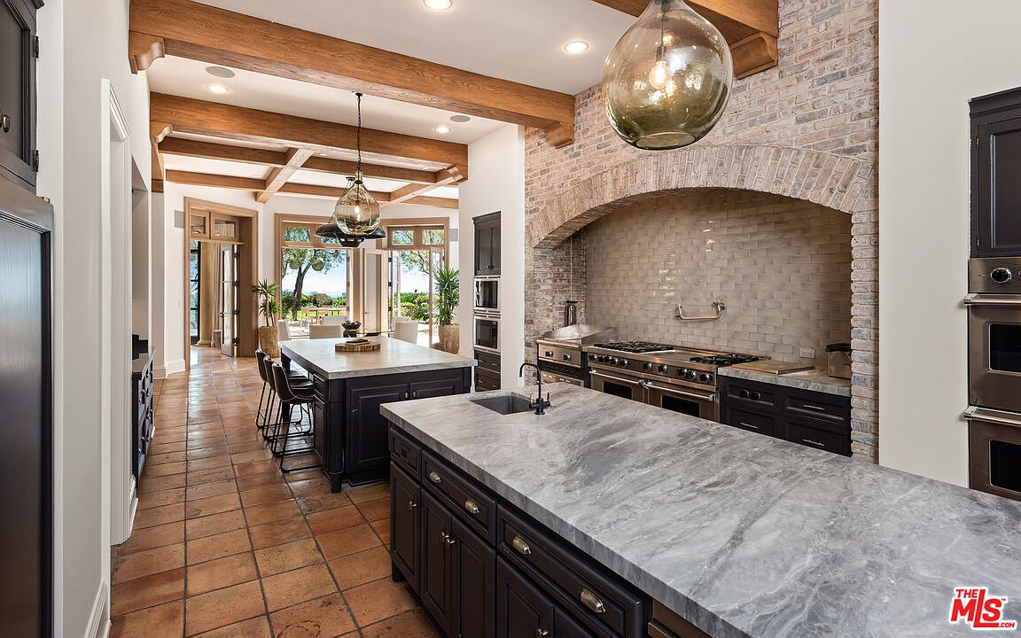 This is a spacious kitchen featuring two islands with marble countertops and dark wood cabinetry. A brick archway frames the cooking area, which includes stainless steel appliances. The room is well-lit with pendant lighting and natural light from the adjacent dining area, creating a warm and inviting atmosphere.