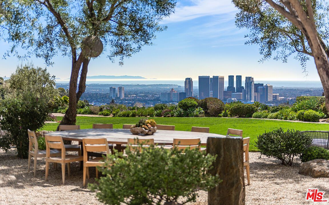 This exterior shot showcases a beautifully landscaped yard with a stunning city view. A large, oval wooden table with chairs sits on a gravel area, suggesting an outdoor dining space. The lush green lawn, manicured bushes, and strategically placed trees create a serene and inviting atmosphere, perfect for entertaining or relaxing while enjoying the panoramic vista.
