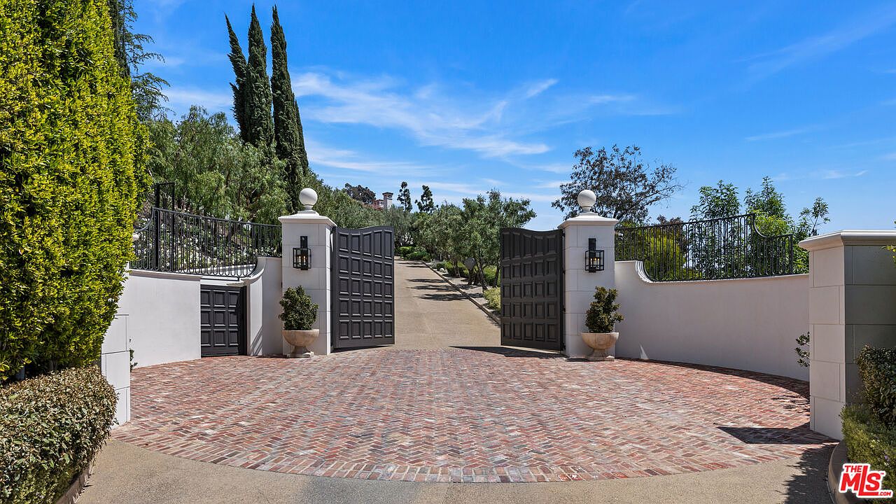 This image showcases an impressive entryway to a property, featuring a brick-paved circular driveway leading to an open, dark-colored gate flanked by white pillars with spherical tops. Lush greenery surrounds the entrance, adding to the sense of privacy and exclusivity. The perspective is from the outside looking in, creating a welcoming and grand impression.