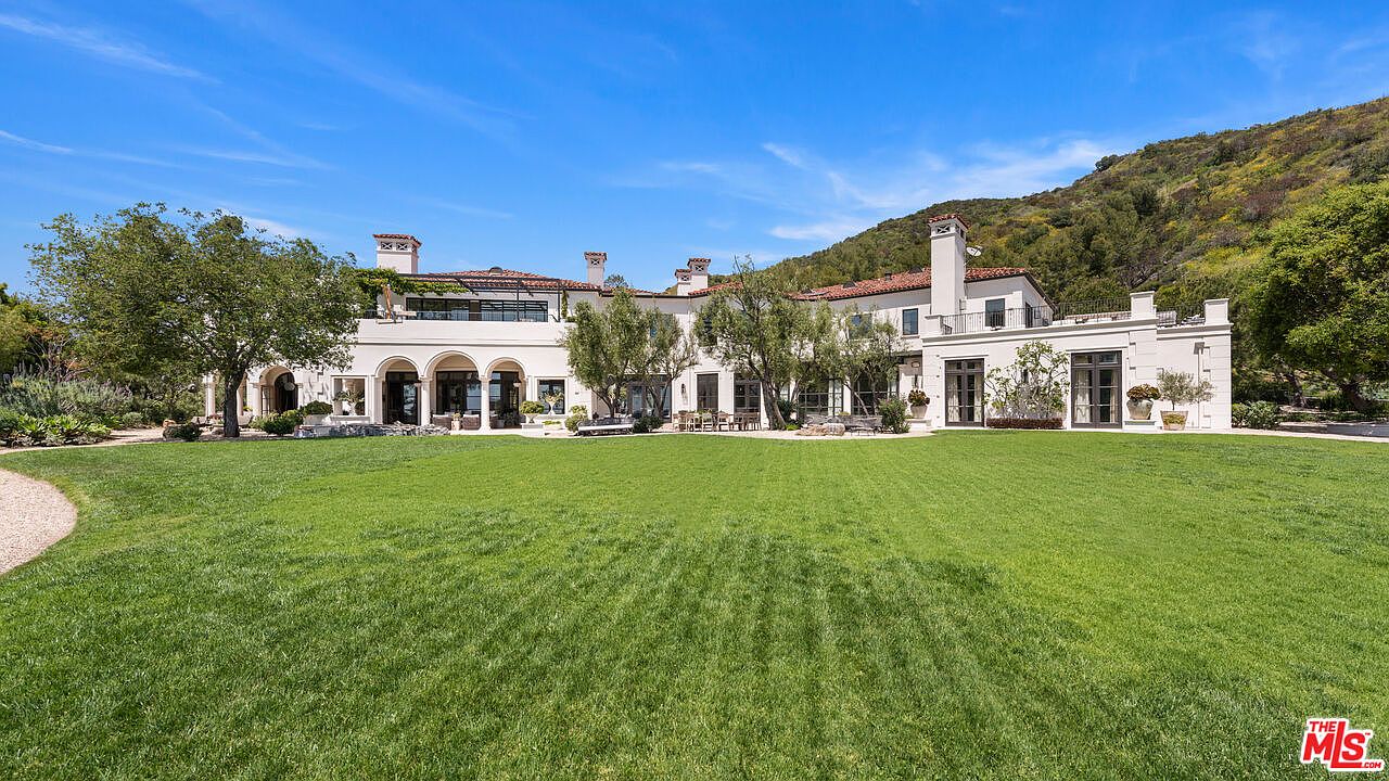 This image showcases the rear exterior of a luxurious estate, featuring a sprawling green lawn that extends to the base of the house. The architecture is elegant with white walls, arched openings, and a red tile roof. The landscape includes mature trees and a hillside backdrop, creating a serene and private setting.