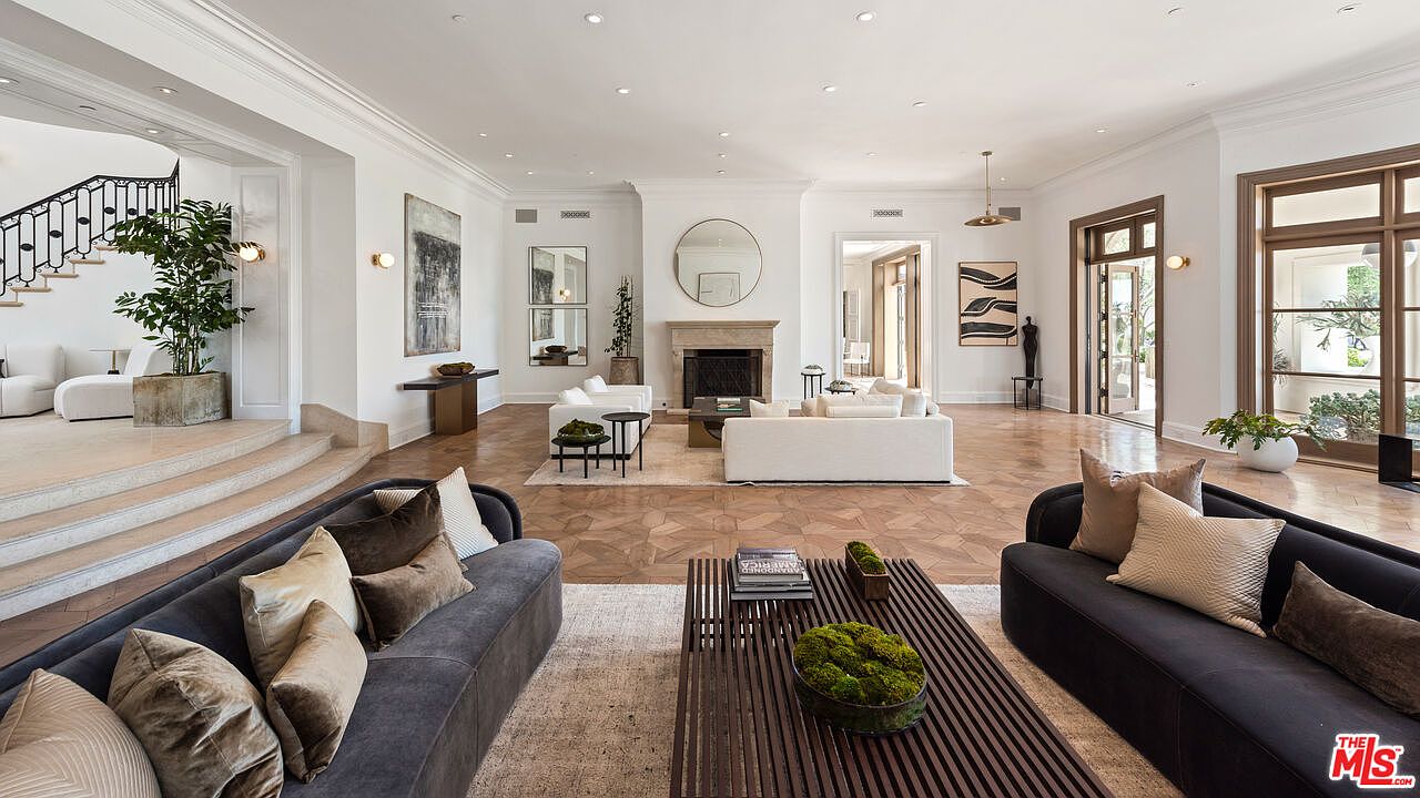 This is a spacious and luxurious living room featuring a neutral color palette with dark accents. The room is well-lit with natural light and recessed lighting, highlighting the hardwood floors and elegant furnishings. Two sofas face each other across a large wooden coffee table, and a fireplace with a round mirror above it serves as a focal point.