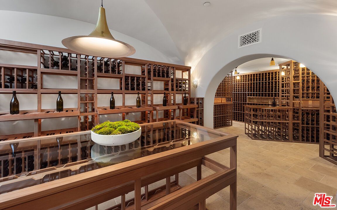 This is an interior shot of a wine cellar featuring extensive wooden wine racks filled with bottles. A large, glass-topped table with a moss centerpiece sits in the foreground, illuminated by a modern pendant light. The room has a vaulted ceiling and an arched doorway leading to another section of the cellar, creating a sophisticated and inviting space for wine storage and tasting.