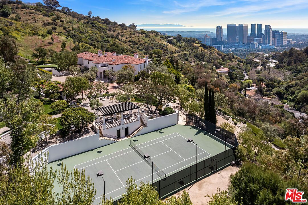 This aerial shot showcases a luxurious estate nestled in a hilly landscape, featuring a grand house with a red tile roof, a tennis court, and a multi-level outdoor structure with a pergola. The property is surrounded by lush greenery, offering privacy and stunning views of the city skyline in the distance. The overall impression is one of opulence and tranquility.