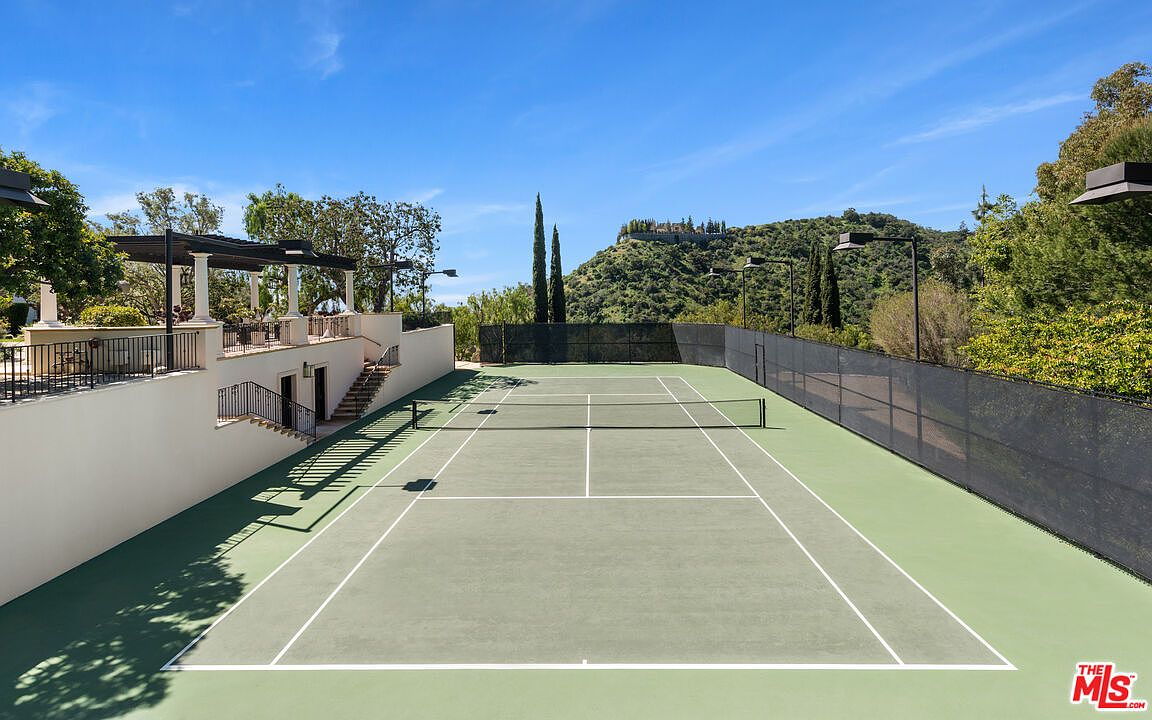 This image showcases a well-maintained tennis court with a green surface and crisp white lines, set against a backdrop of lush greenery and a clear blue sky. A building with a covered patio overlooks the court, adding a touch of luxury and providing a viewing area. The scene conveys a sense of recreation and upscale outdoor living.