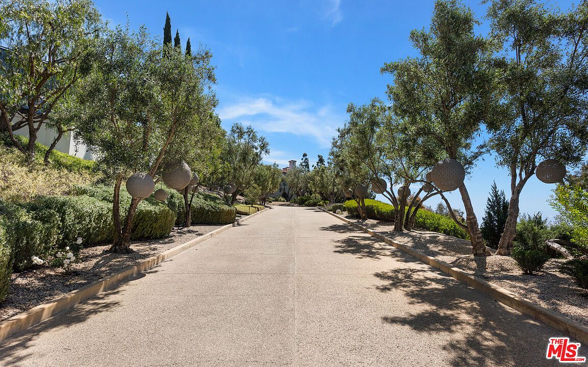 This image showcases a grand entryway leading to a luxurious property. The long, paved driveway is lined with meticulously maintained trees and shrubbery, creating a sense of privacy and elegance. Decorative spherical elements hang from the trees, adding a unique touch to the landscape design, while the clear blue sky enhances the overall appeal.