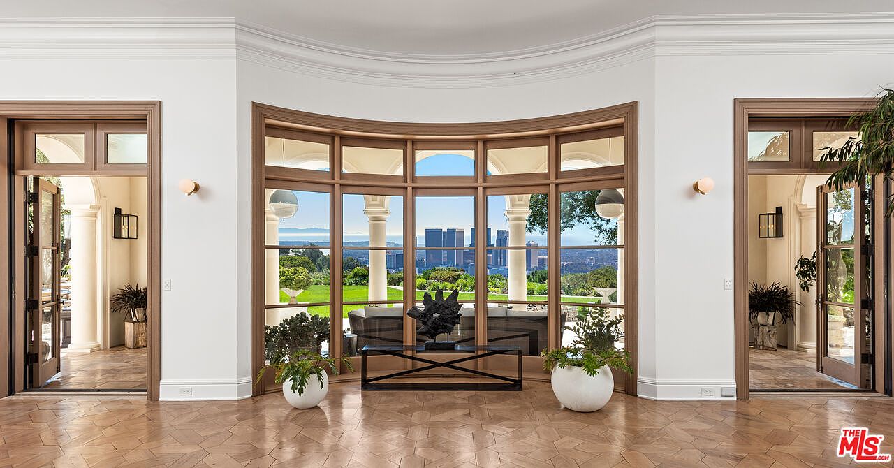 This interior shot showcases a grand hallway with herringbone wood flooring, flanked by doorways and a large, curved window offering a stunning city view. The space is decorated with potted plants and a modern sculpture on a console table, creating an elegant and inviting atmosphere. The architectural details, including the crown molding and paneled doors, add to the luxurious feel of the home.