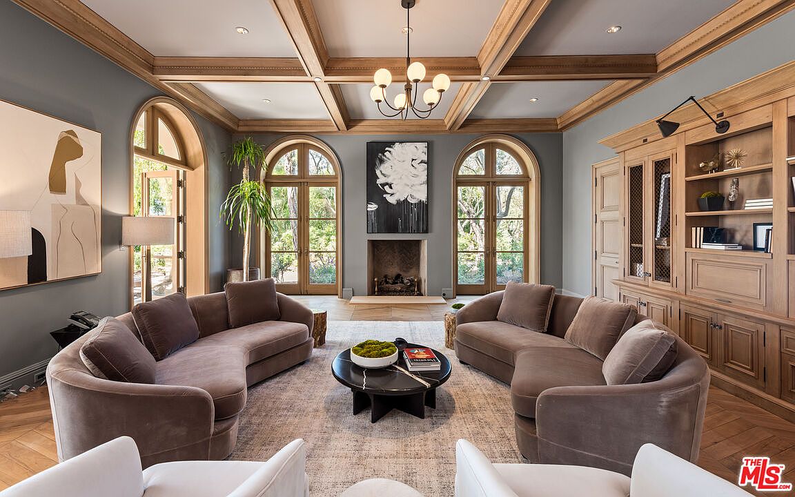 This is an interior shot of a luxurious living room featuring two curved sofas facing each other, centered around a round coffee table. The room has a sophisticated color palette with gray walls, light wood trim, and herringbone wood floors. Arched windows and a fireplace with a black and white painting above it add architectural interest, while a built-in cabinet provides ample storage.