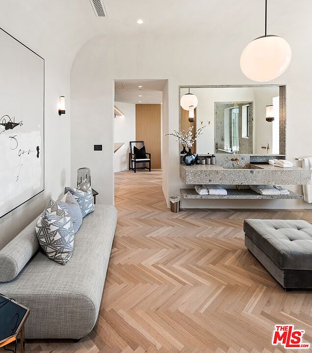This is a luxurious primary bathroom featuring a modern design with a herringbone wood floor and a floating vanity with a stone countertop. A large mirror reflects the light from the pendant light, enhancing the bright and airy feel of the space. A comfortable lounge area with a sofa and ottoman adds a touch of relaxation to the bathroom.