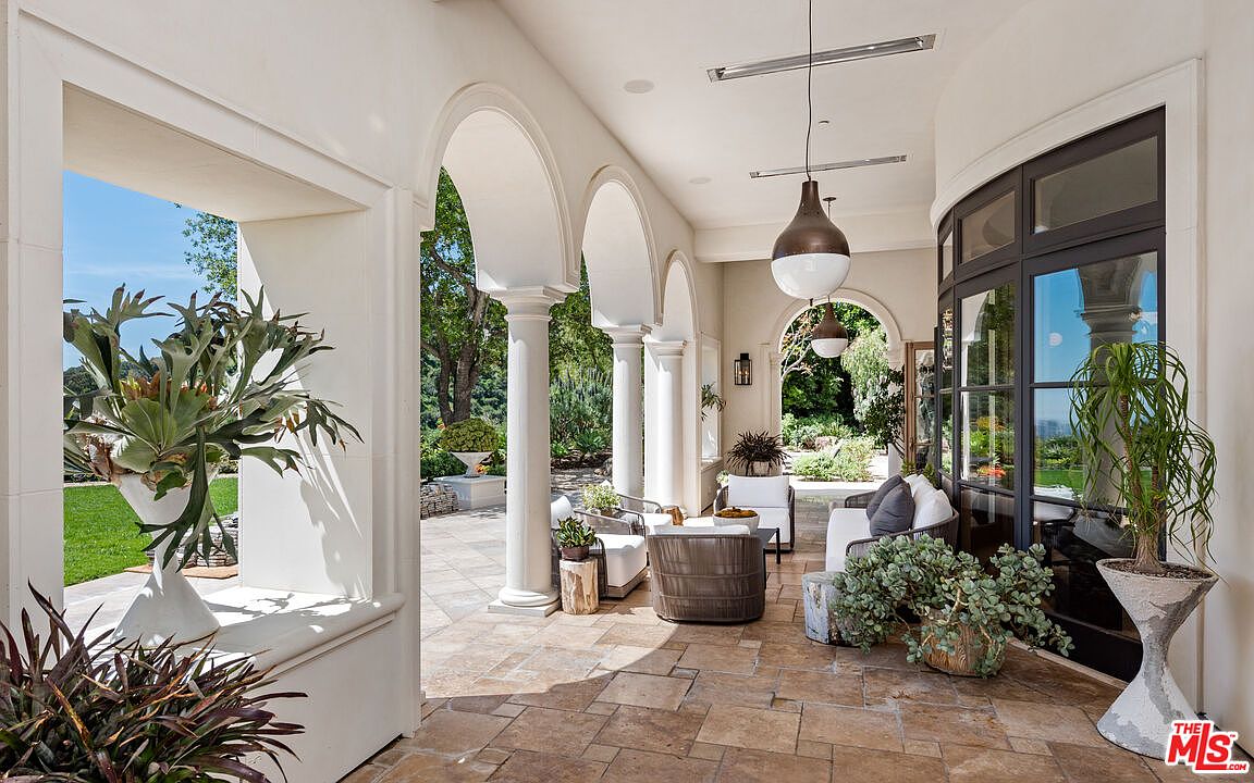 This image showcases a luxurious patio or balcony area, featuring elegant architectural details such as arched openings and columns. The space is furnished with comfortable seating arrangements and decorated with lush greenery, creating a serene and inviting atmosphere. The flooring is tiled, and the overall design exudes sophistication and relaxation.