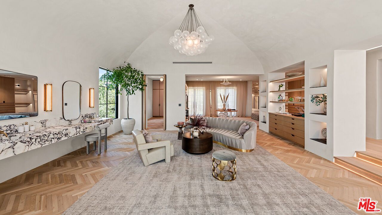 This is a spacious and luxurious living room featuring a curved sofa, a round coffee table, and a stylish accent chair. The room is well-lit with a modern chandelier and natural light from a large window. The herringbone wood floors and a large area rug add warmth and texture to the space, creating an inviting and sophisticated atmosphere.