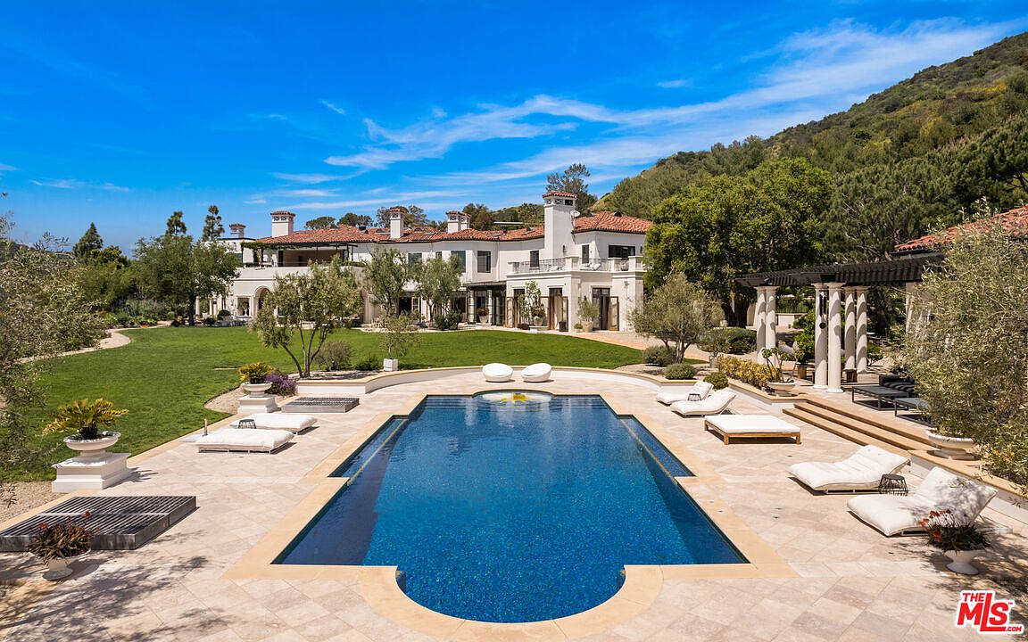 This image showcases a luxurious backyard featuring a rectangular pool with a dark blue interior and light-colored stone surround. Several lounge chairs are arranged around the pool, inviting relaxation. The backdrop includes a large, elegant house with a red tile roof, a well-manicured lawn, and lush greenery, creating a serene and upscale atmosphere.