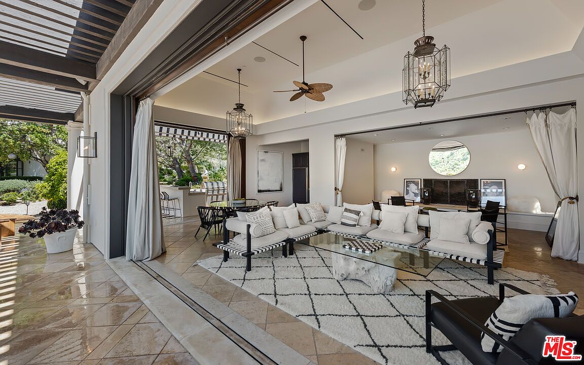 This is an interior shot of a luxurious living room that seamlessly blends indoor and outdoor spaces. The room features a stylish seating arrangement with a modern sofa, a glass coffee table, and a patterned rug. Large sliding doors open to an outdoor patio, enhancing the sense of spaciousness and natural light. The color palette is neutral with black accents, creating a sophisticated and inviting atmosphere.
