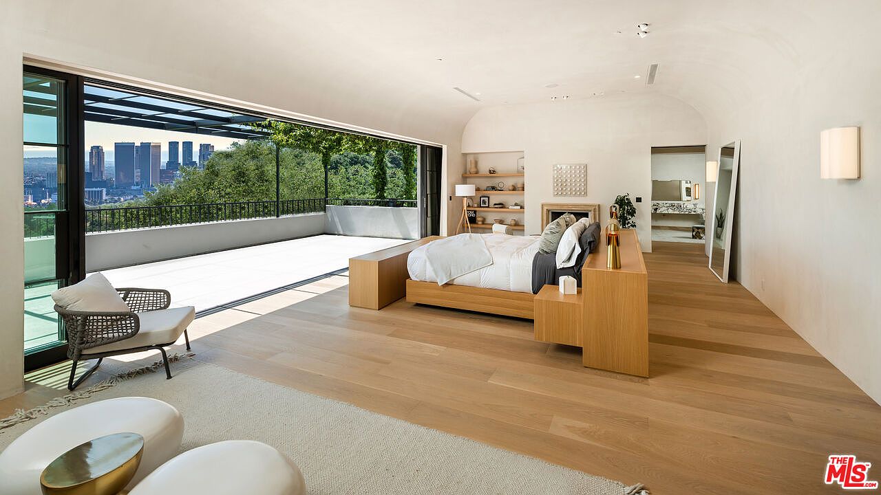 This is a spacious primary bedroom featuring a modern design with light wood flooring and a minimalist aesthetic. The room opens onto a large balcony with city views, creating an indoor-outdoor living experience. The bed is a platform style with integrated side tables, and the room includes a seating area with modern chairs and coffee tables.