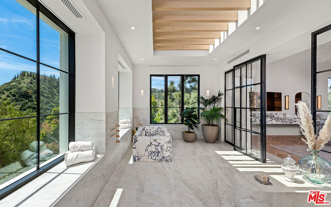 This is a luxurious primary bathroom featuring marble flooring and walls, a freestanding marble bathtub, and large windows offering scenic views. The bathroom includes modern fixtures, a glass-enclosed vanity area, and stylish decor, creating a spa-like atmosphere. The room is bright and spacious, emphasizing high-end design and comfort.