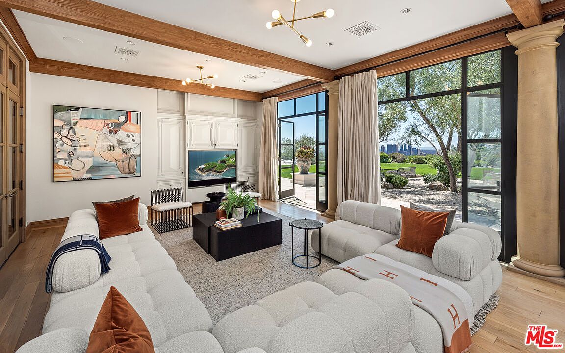 This is a luxurious living room featuring a large, modular white sofa with rust-colored pillows, a dark coffee table with plants, and a view of the outdoor landscape through expansive windows. The room is accented with wooden beams, built-in cabinetry, and modern lighting fixtures, creating a sophisticated and comfortable atmosphere. The perspective is from the corner of the room, capturing the depth and breadth of the space.