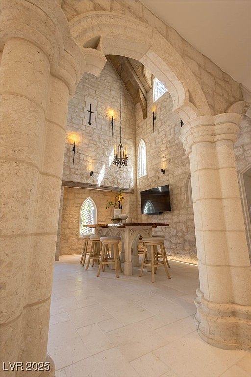 This interior shot showcases a unique dining area with prominent architectural features, including stone columns and arched openings. A high table with bar stools sits in the center, illuminated by a chandelier, while a wall-mounted television adds a modern touch. The room's design evokes a sense of historical grandeur blended with contemporary living.