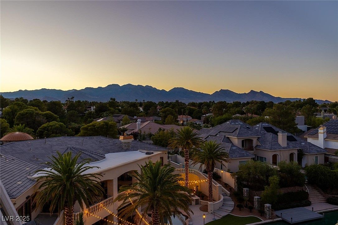 This aerial view showcases a luxurious residential property at dusk, featuring multiple buildings with tiled roofs and well-manicured landscaping. Palm trees adorned with string lights add a touch of elegance, while the backdrop of distant mountains enhances the scenic appeal. The overall impression is one of upscale living and serene beauty.