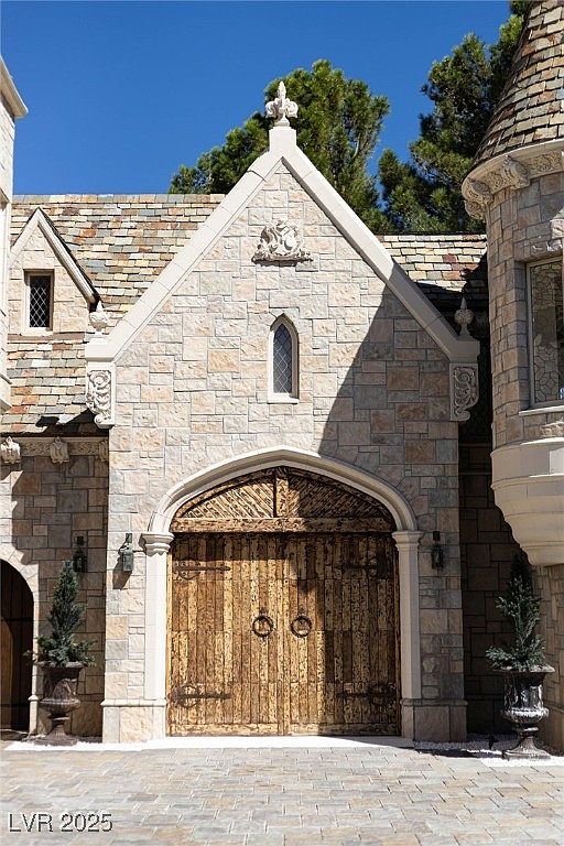 The image showcases a grand, castle-like garage exterior. The structure is built with light-colored stone, featuring an arched wooden door with metal accents and ring handles. The architectural style evokes a sense of old-world charm and luxury, complemented by manicured greenery on either side and a stone-paved driveway.