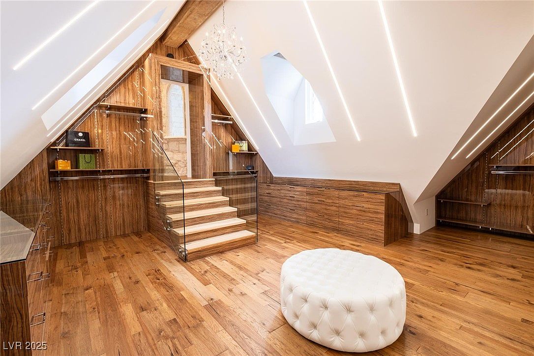 This is an interior shot of a luxurious walk-in closet featuring wood cabinetry, a staircase leading to an upper level, and a chandelier. The closet is well-lit with modern lighting fixtures and includes a tufted ottoman in the center. The overall impression is one of high-end design and ample storage space.