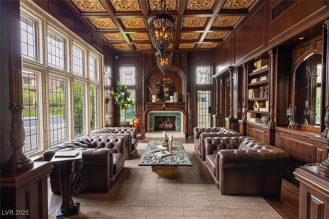 This is an opulent living room featuring dark wood paneling, an intricately designed coffered ceiling, and two ornate chandeliers. Two sets of tufted leather sofas face each other, flanking a decorative coffee table and fireplace. Large windows provide natural light, creating a luxurious and inviting atmosphere.
