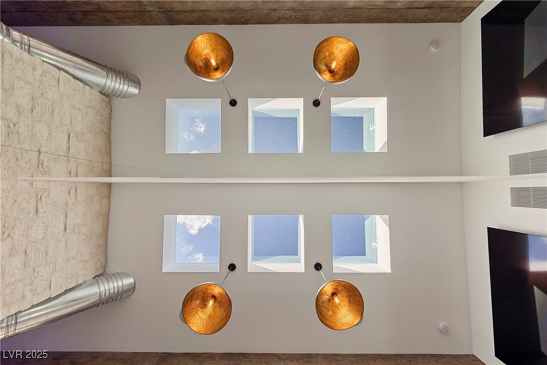 This image showcases a unique architectural detail, featuring a ceiling with multiple square skylights that allow natural light to flood the space. Modern pendant lights hang below the skylights, adding an element of contemporary design. The walls are a neutral color, and there's a textured wall on the left side, creating a visually interesting contrast.