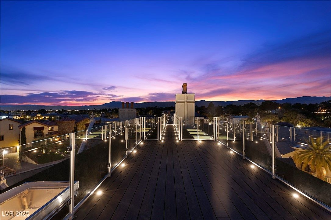 This image showcases a modern rooftop deck at dusk, featuring sleek, dark-toned decking and glass railings with integrated lighting. The deck offers panoramic views of the surrounding cityscape and distant mountains under a vibrant, colorful sky. The overall impression is one of luxury and sophisticated outdoor living.