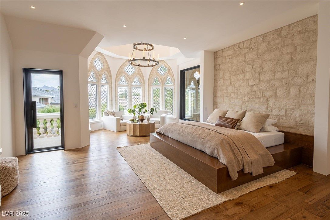 This is a luxurious primary bedroom featuring a modern platform bed with a neutral-toned duvet and multiple pillows. The room boasts unique architectural details, including arched windows with intricate lattice work and a stone accent wall. The hardwood flooring and area rug add warmth and texture to the space, creating an inviting and sophisticated atmosphere.
