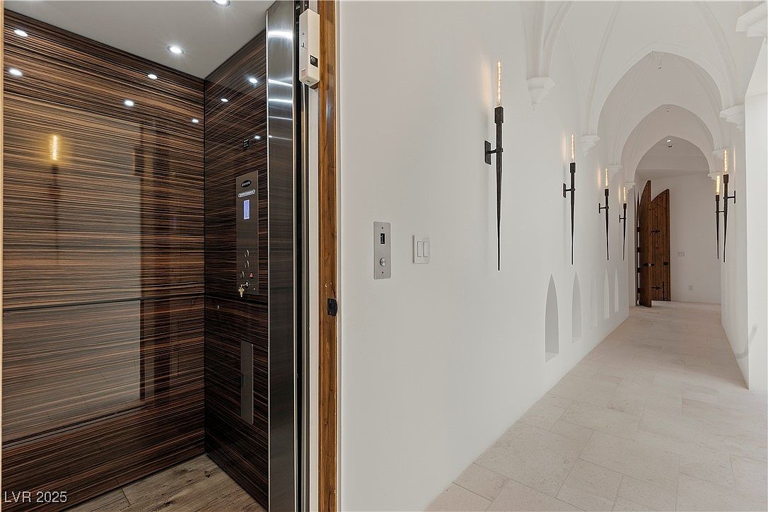 This interior shot showcases a modern hallway with unique architectural details. An elevator with wood paneling and stainless steel accents is visible on the left, while the hallway features arched ceilings, sconce lighting resembling torches, and tiled flooring. The overall impression is a blend of modern convenience and gothic-inspired design.