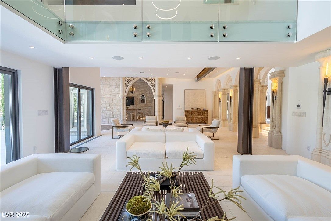 This is a spacious and luxurious living room featuring white sofas, a modern coffee table with decorative plants, and elegant architectural details such as stone arches and columns. The room is well-lit with natural light and recessed lighting, creating a bright and inviting atmosphere. The design incorporates a blend of modern and classic elements, making it a sophisticated space for relaxation and entertainment.